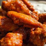 A close-up of shiny, saucy Extra Crispy Baked Buffalo Chicken Wings piled high, with blue cheese dressing visible in the background.