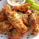 A plate of crispy, golden brown Garlic Parmesan Air Fryer Chicken Wings topped with grated cheese and parsley, served with celery sticks and dip.