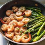 Close-up of shrimp and asparagus cooking in a skillet, seasoned and topped with grated cheese.