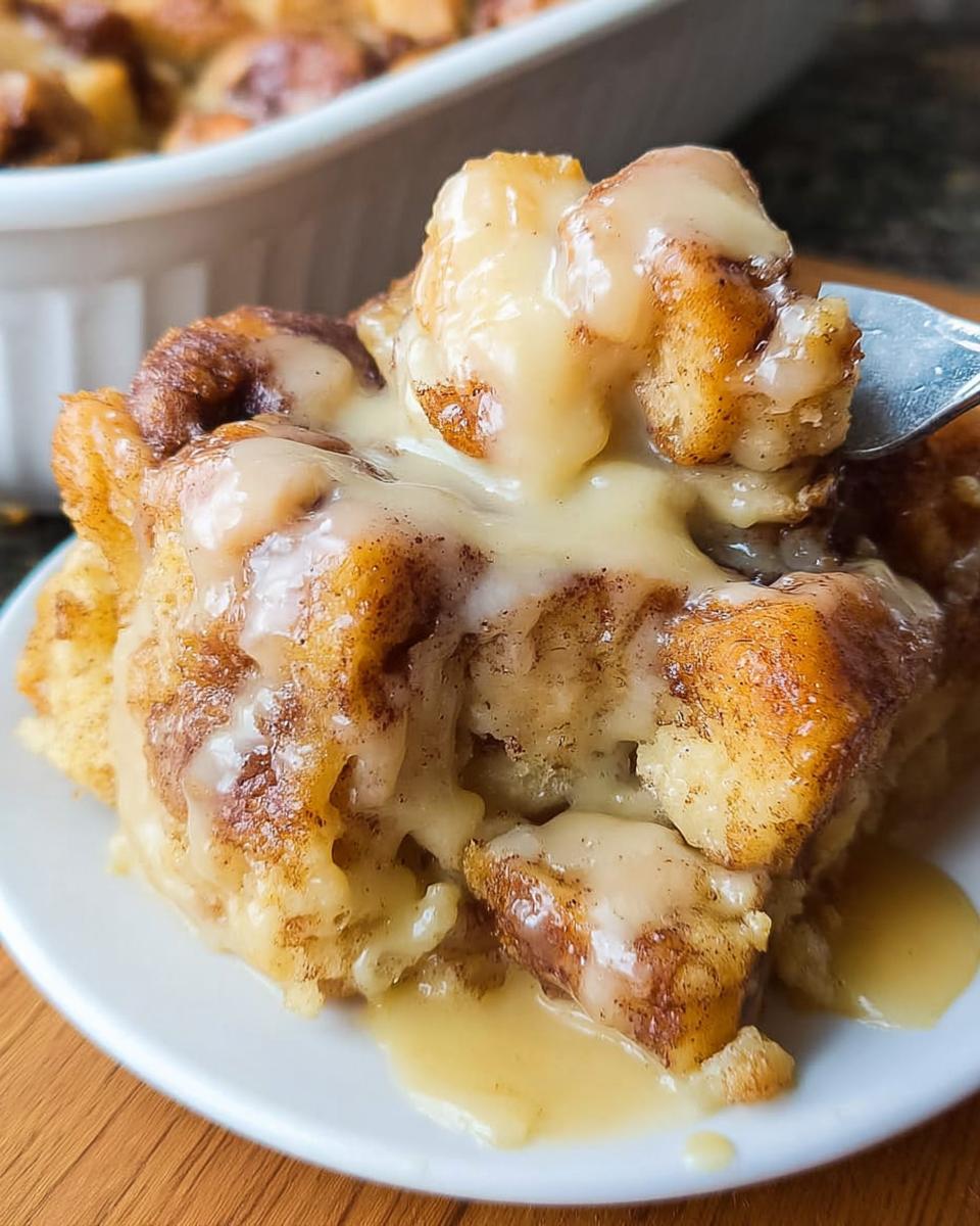 A fork lifts a piece of Gooey Cinnamon Roll Bread Pudding covered in thick vanilla icing.