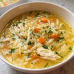 A close-up view of a bowl of vibrant Lemon Orzo Chicken Soup, featuring shredded chicken, carrots, orzo pasta, and parsley garnish.