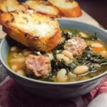 A bowl of Rustic Tuscan White Bean and Kale Soup topped with sausage and a piece of cheesy toasted bread.