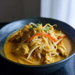 A close-up of a dark bowl filled with Spicy Thai Coconut Curry Noodle Soup, featuring yellow curry broth, noodles, chicken, and bean sprouts.