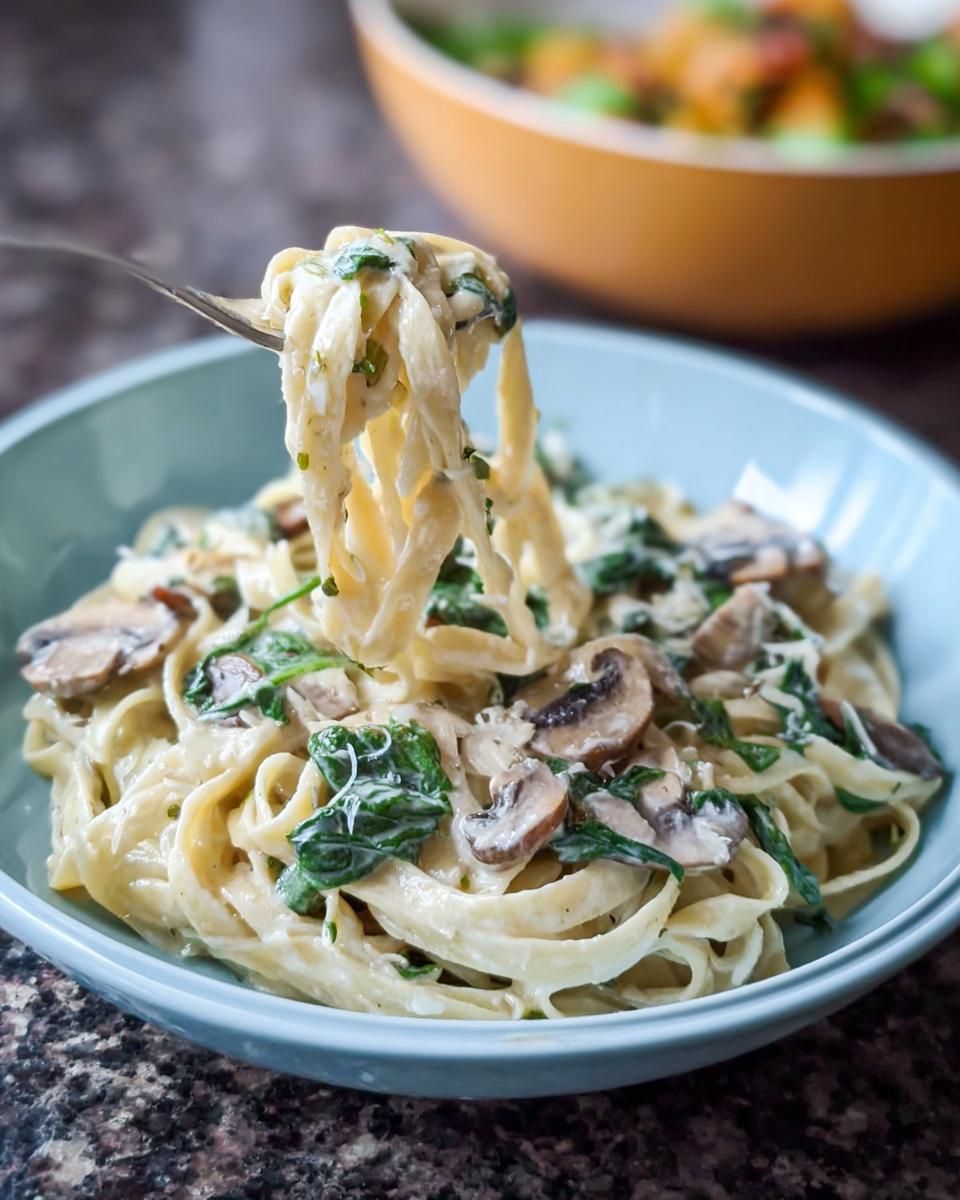 A fork lifting fettuccine noodles coated in creamy sauce from a bowl of Spinach and Mushroom Alfredo Pasta.