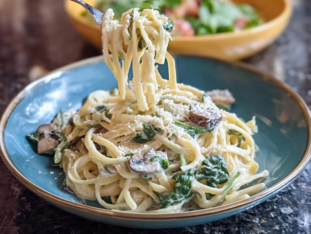 A fork lifts creamy fettuccine noodles coated in sauce, spinach, and mushrooms from a blue bowl of Spinach and Mushroom Alfredo Pasta.