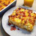 A square slice of cheesy bacon and egg breakfast casserole on a white plate, ready to eat.