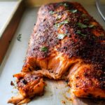 Close-up of a large, perfectly baked salmon fillet with a dark spice rub, flaking easily on a metal baking sheet.