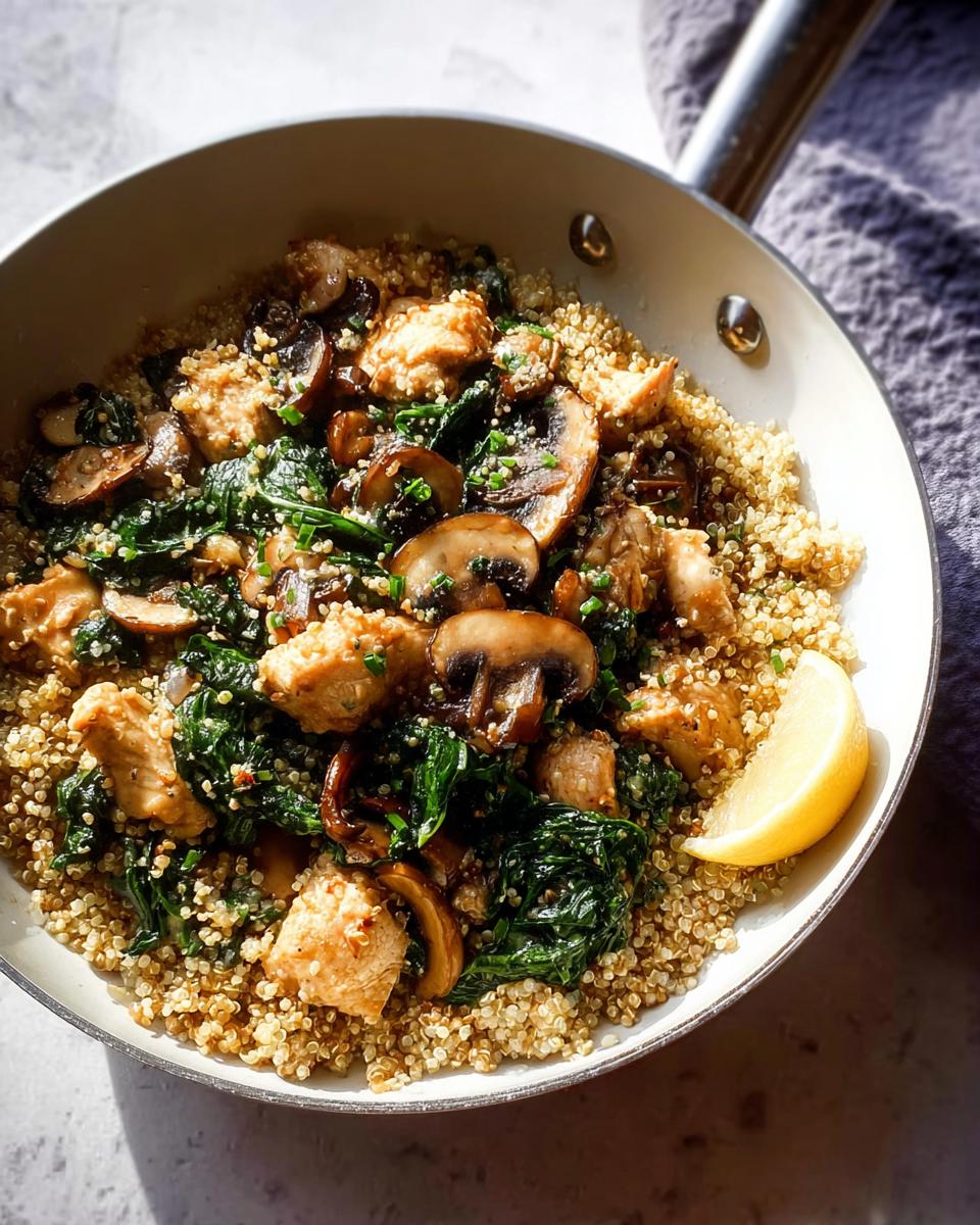 A skillet meal featuring small pieces of chicken, sautéed mushrooms, spinach, and quinoa, garnished with a lemon wedge.