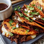 Close-up of cheesy birria tacos topped with cilantro and onion, served with a side of consommé for The Ultimate Taco Tuesday Recipes Guide.
