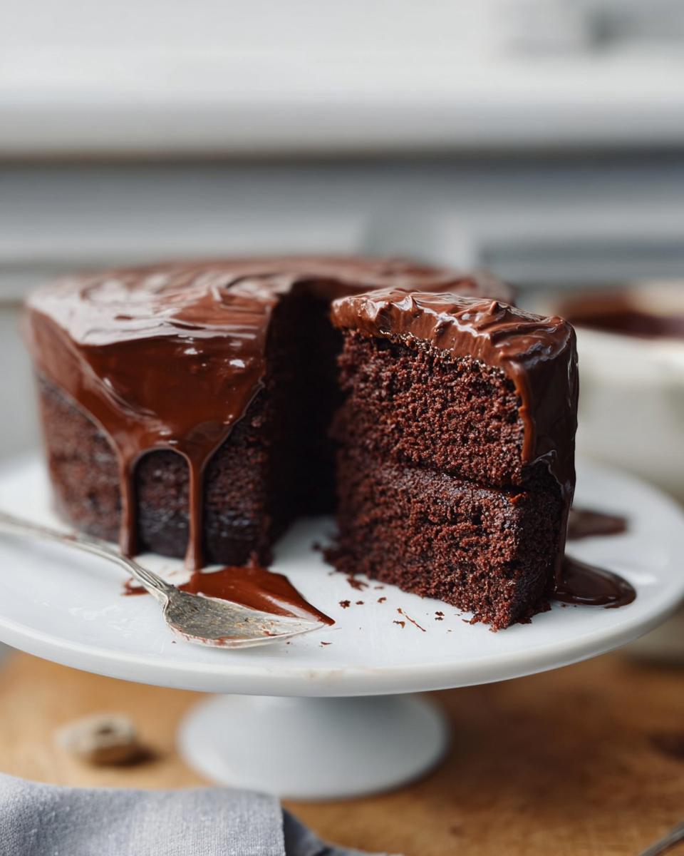 A rich, two-layer chocolate cake with thick, glossy ganache frosting, featuring a slice cut out. A perfect example of 7-Ingredient Cake Ideas.