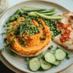 Bright orange hummus topped with green peas and fresh herbs, served with pita bread and cucumber slices, perfect for easy dinner recipes.
