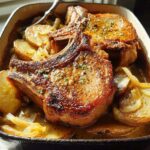 Two perfectly seared bone-in pork chops resting on a bed of sliced potatoes and onions in a baking dish.