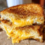 Close-up of a perfectly golden brown grilled cheese sandwich cut in half, showing melted cheddar cheese oozing out.