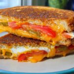 A stack of two halves of a grilled cheese sandwich overflowing with melted cheddar, tomato slices, and jalapeños, part of our 20-Ingredient Grilled Cheese Recipes.