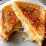 Close-up of a perfectly golden brown grilled cheese recipe, cut in half, showing a dramatic, gooey cheese pull.