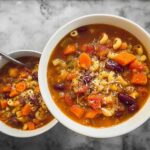 Two bowls of hearty vegetable soup recipes, full of beans, carrots, pasta, and topped with cheese.