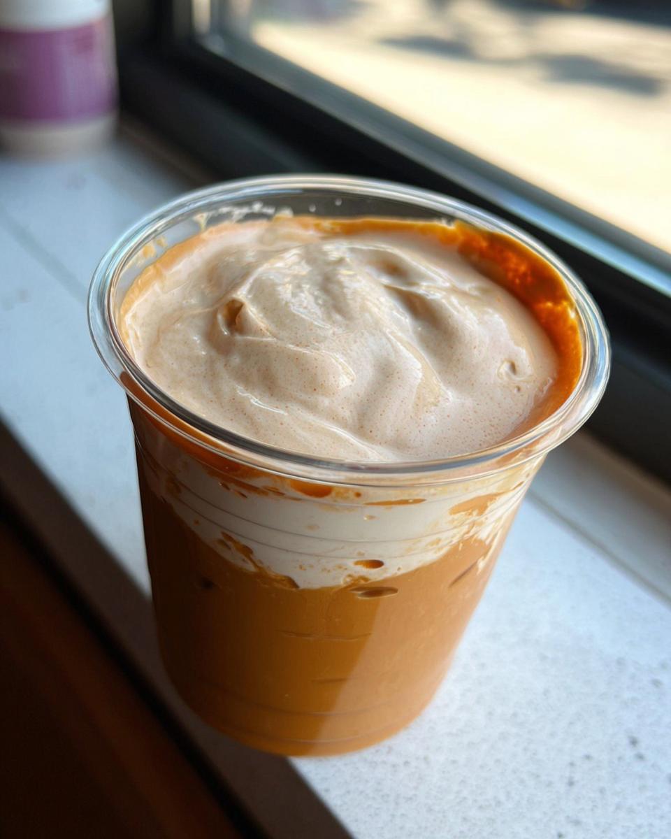 Close-up of an iced orange-colored beverage in a clear plastic cup topped with thick, creamy foam, resembling Restaurant-Style Starbucks Drinks Recipes at Home.