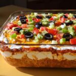 Close-up of a layered taco dip in a glass dish, topped with cheese, olives, tomatoes, and green onions.