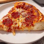 A thick slice of pepperoni pizza with melted, stretching cheese sits on a white plate, perfect for Pizza Night Recipes Meal Prep.