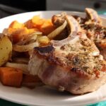 Two bone-in pork chops recipes seasoned with herbs served alongside roasted potatoes, carrots, and onions on a white plate.