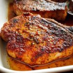 Close-up of a thick, perfectly seared pork chop with a rich, seasoned crust resting in pan juices.