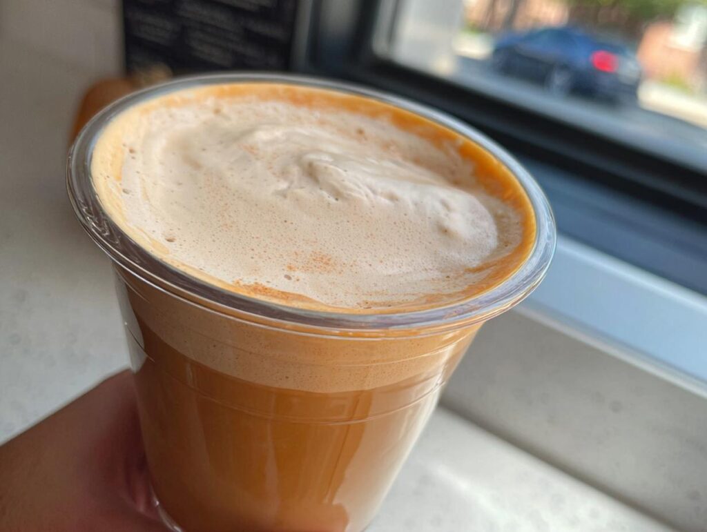 Close-up of a hand holding a cold, orange-colored drink with thick foam, resembling Restaurant-Style Starbucks Drinks Recipes at Home.
