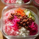 Close-up of a glass container holding one of the Rice Bowls Recipes Meal Prep That Actually Tastes Great, featuring pink rice, seasoned ground meat, pickled onions, and cucumbers.