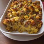 A close-up of a baked sausage cornbread stuffing in a white casserole dish, one serving removed.