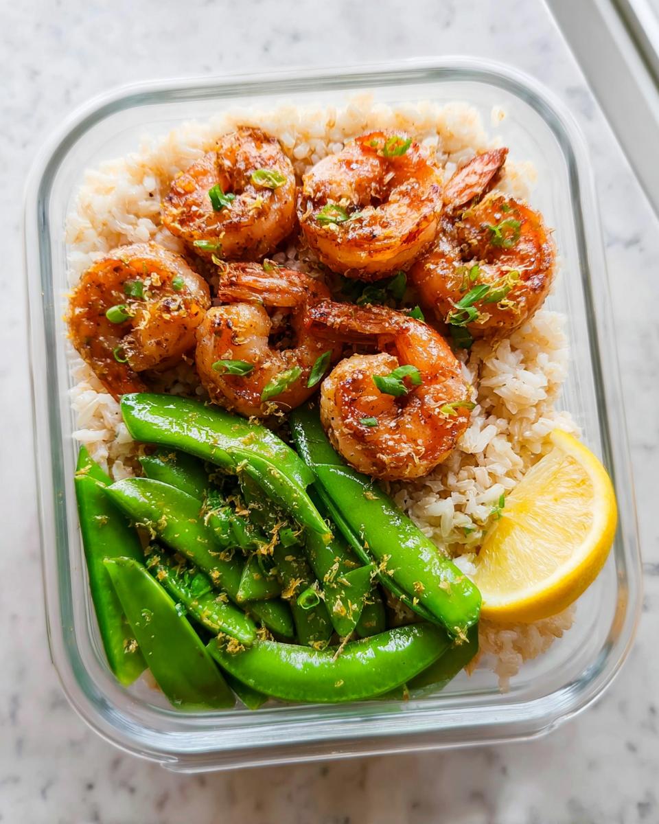 Overhead view of a glass meal prep container filled with brown rice, seasoned shrimp, and bright green snap peas, garnished with lemon zest.