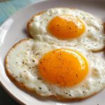 Two sunny side up fried eggs, seasoned with black pepper, served on a white plate. These are great egg recipes.