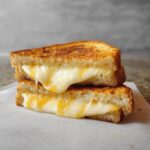 Two halves of a perfectly golden brown grilled cheese sandwich stacked, showing gooey, melted cheese oozing out.