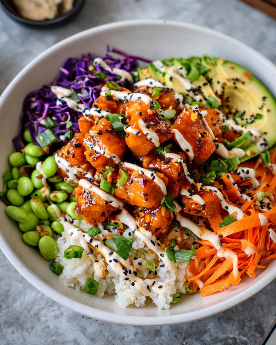Close-up of a bowl filled with rice, crispy Bang Bang Salmon Bites, avocado slices, shredded carrots, edamame, and drizzled with sauce.