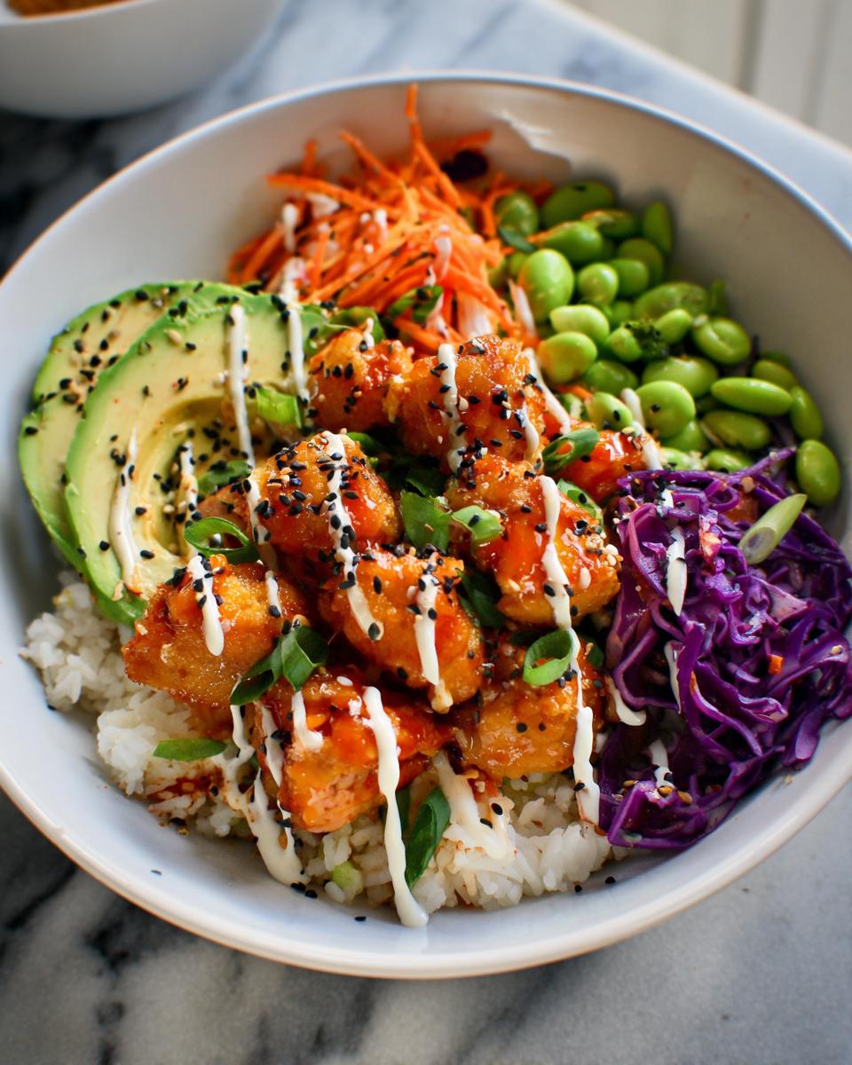 Close-up of a bowl featuring crispy Bang Bang Salmon Bites over rice, topped with avocado, edamame, carrots, and a drizzle of white sauce.