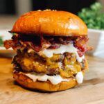 A tall, juicy double patty Crack Burger loaded with melted cheese, crispy bacon, and white sauce on a brioche bun.