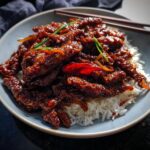 A close-up of Crispy Chilli Beef Rice, featuring dark, glossy beef strips over white rice, garnished with spring onions.