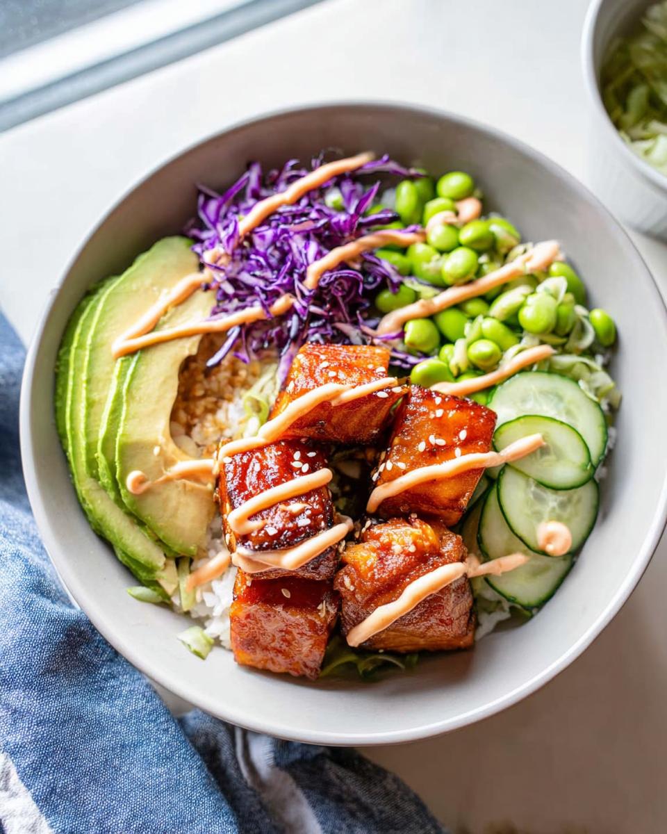 Close-up of a Crispy Salmon and Rice Bowl featuring glazed salmon cubes, avocado slices, edamame, and purple cabbage with a drizzle of sauce.