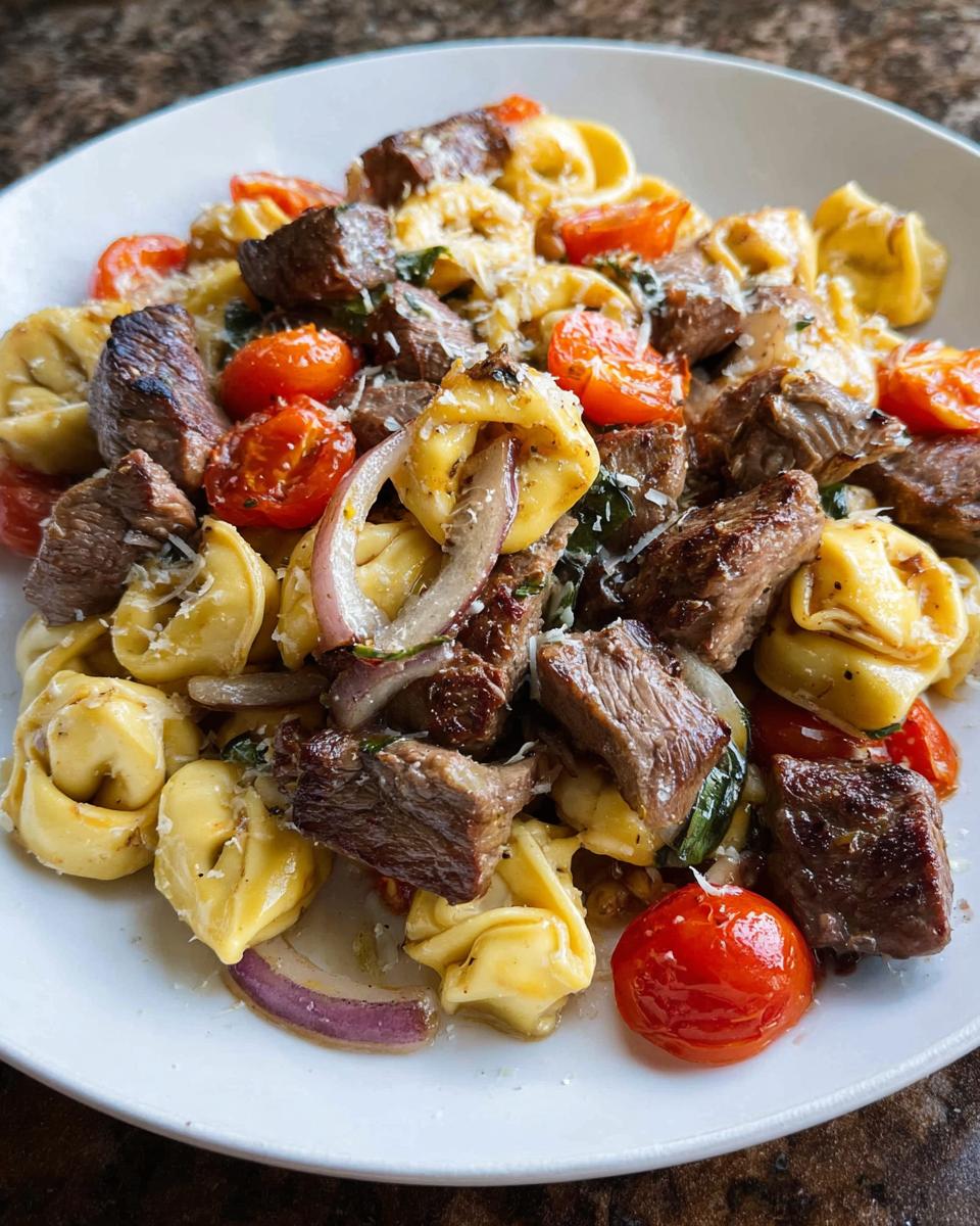A close-up of a white bowl filled with Garlic Steak Tortellini, featuring chunks of seared steak, cherry tomatoes, and red onion.