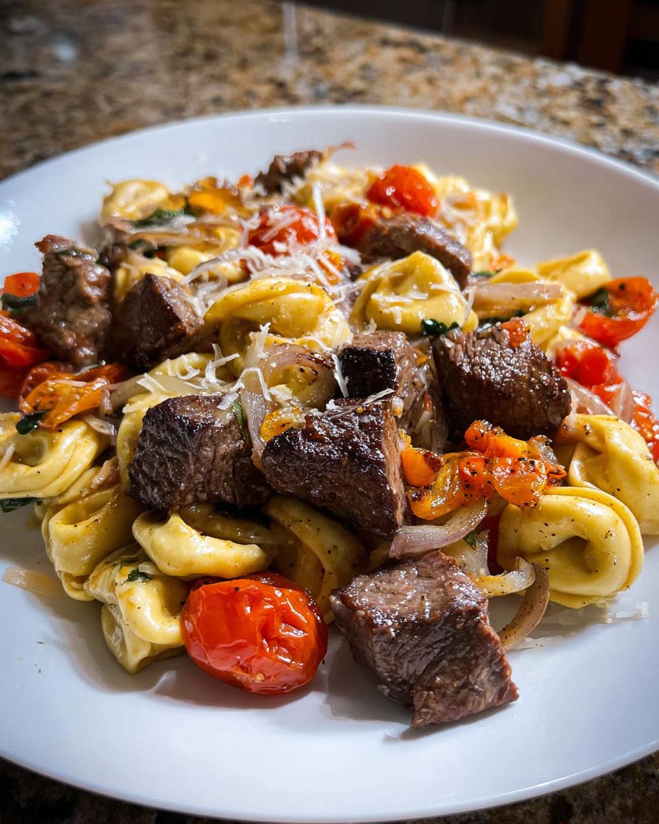 Close-up of a white bowl filled with Garlic Steak Tortellini, featuring chunks of seared steak, roasted cherry tomatoes, and shaved Parmesan.