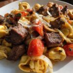 A close-up view of a serving of Garlic Steak Tortellini mixed with roasted tomatoes and onions, topped with shaved cheese.