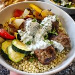 A close-up of a Greek Meatball Bowl Recipe featuring meatballs, roasted vegetables, Israeli couscous, and tzatziki sauce.