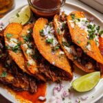 A plate featuring four crispy, red-orange My Fave Birria Tacos topped with cheese, onions, and cilantro, served with lime and consommé.
