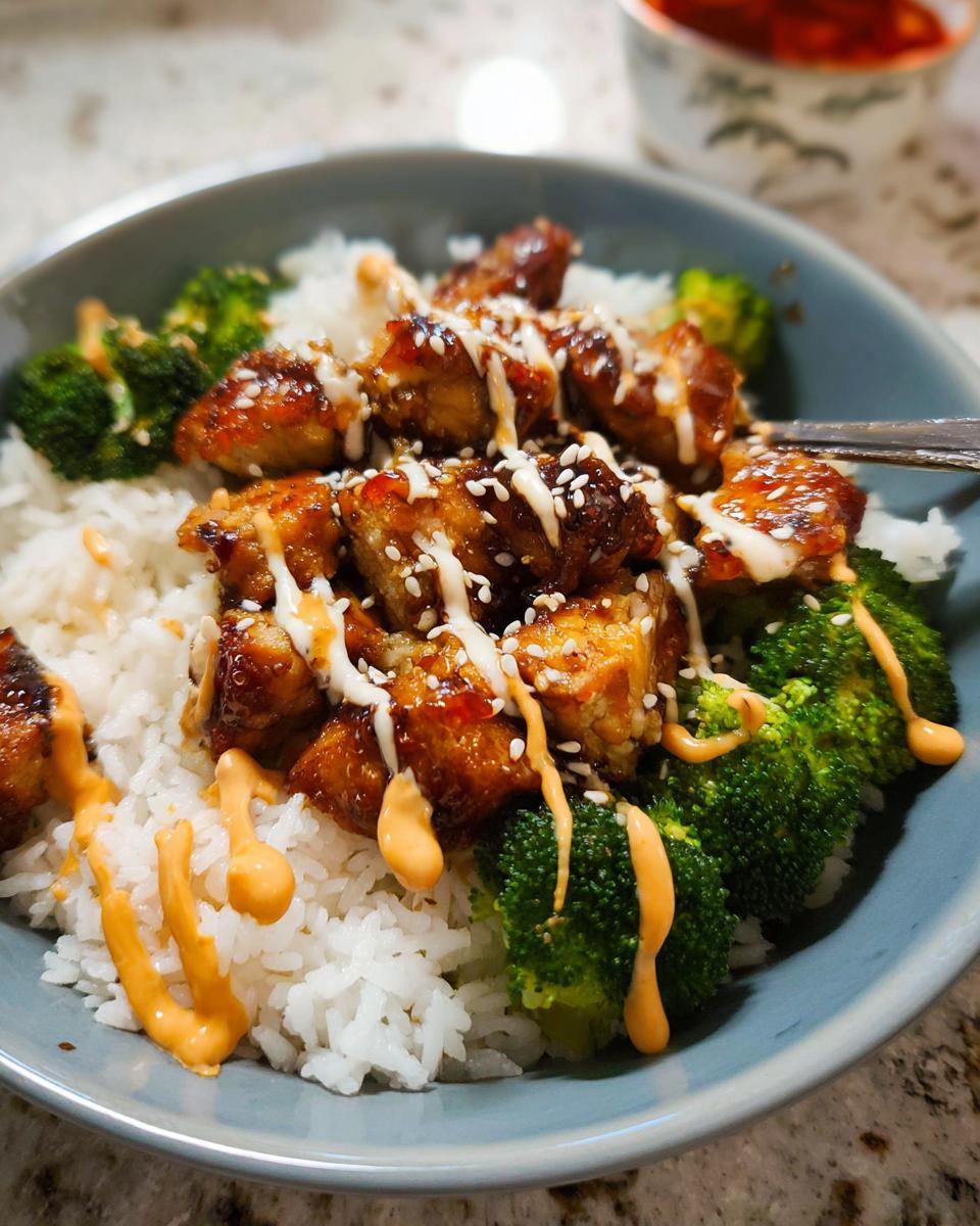 A close-up of a serving of Sticky Chicken Bowls featuring glazed chicken pieces over white rice and broccoli, drizzled with sauces and sesame seeds.