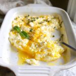 A close-up of baked feta eggs in a white baking dish, topped with fresh basil and herbs.