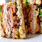 A tall and delicious Breakfast Club Sandwich, cut in half, showing layers of turkey, bacon, cheese, tomato, and lettuce on toasted bread.