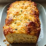 A golden brown cheesy garlic bread loaf with herbs on a white platter.