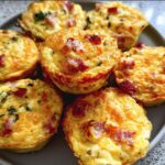 A plate of golden-brown Cheesy Muffin Tin Cottage Cheese Egg Bites with visible pieces of bacon and herbs.