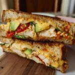 A close-up of a halved Chicken Avocado Melt Sandwich, showing shredded chicken, avocado slices, melted cheese, and red peppers.