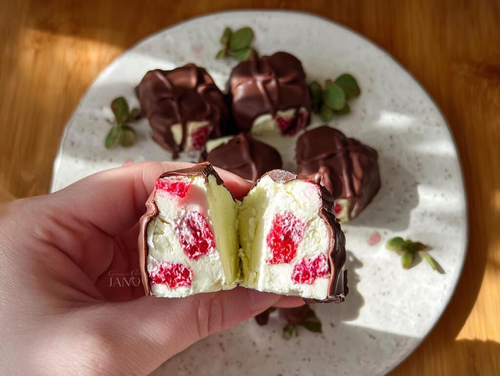 A hand holding a cut Chocolate Strawberry Yogurt Cluster, revealing creamy yogurt and strawberry pieces inside dark chocolate coating.