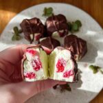 A hand holding a cut Chocolate Strawberry Yogurt Cluster, revealing creamy yogurt and strawberry pieces inside dark chocolate coating.