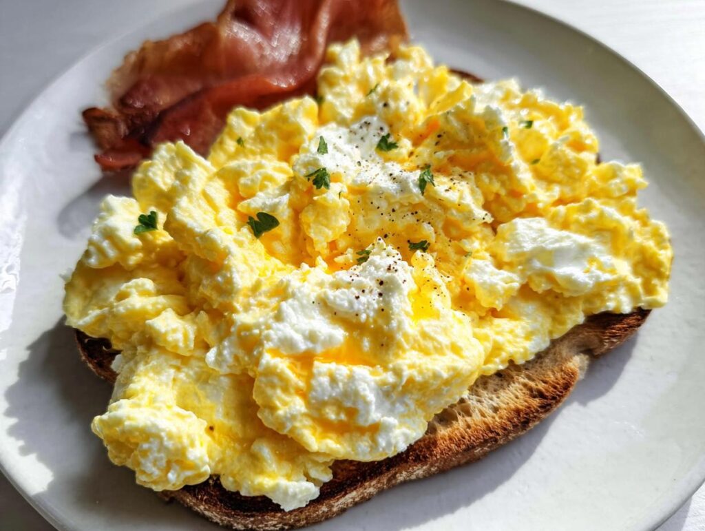 A slice of toast topped with creamy ricotta eggs and a side of bacon. This is Creamy Ricotta Egg Toast.